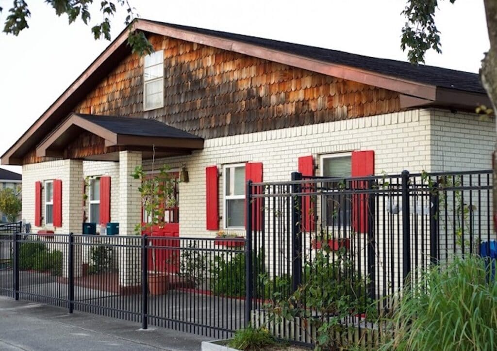 Single-story house with light brick walls, red shutters, wooden shingles on the gable, a black metal fence in front, and some greenery along the entrance—perfect for a Montessori preschool Metairie families trust for a warm and inviting learning environment.
