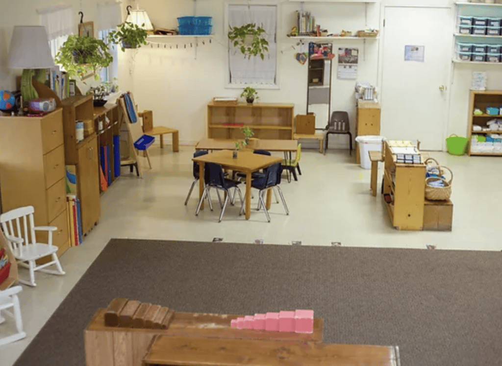 Kinder Haus classroom with wooden furniture, shelves of books and supplies, small tables with chairs, plants, and a carpeted area in the foreground—this warm environment reflects the inviting atmosphere of a Montessori preschool in Metairie.