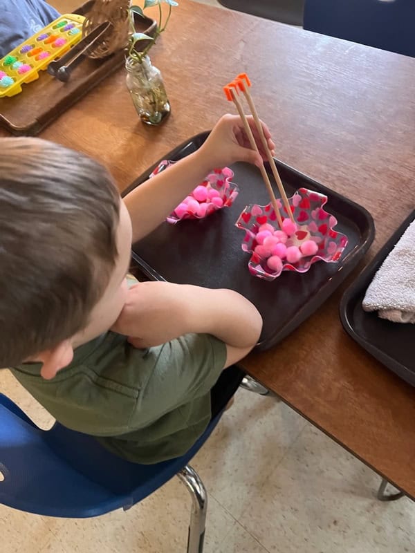 A child uses chopsticks to move pink pom-poms from one polka-dotted bowl to another while seated at a table.