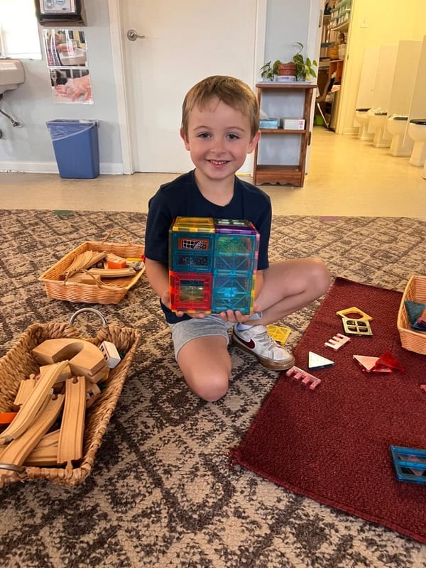 montessori-kindergardner Young boy sitting on a rug in a classroom, smiling and holding a colorful structure made from magnetic tiles, with wooden blocks and more tiles beside him.
