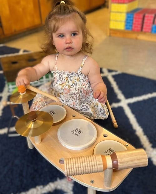 A young child in a floral dress plays a wooden toy drum set with sticks, standing on a blue and white patterned rug.