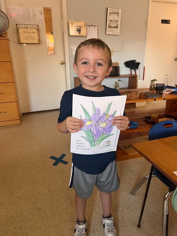 Young boy standing in a classroom holding a colored drawing of a purple flower, smiling at the camera.