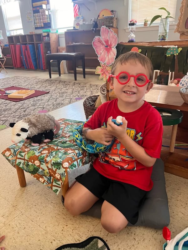 Young boy in red shirt and toy glasses sits on a cushion in a classroom, holding toys, with a stuffed panda on a small table beside him.
