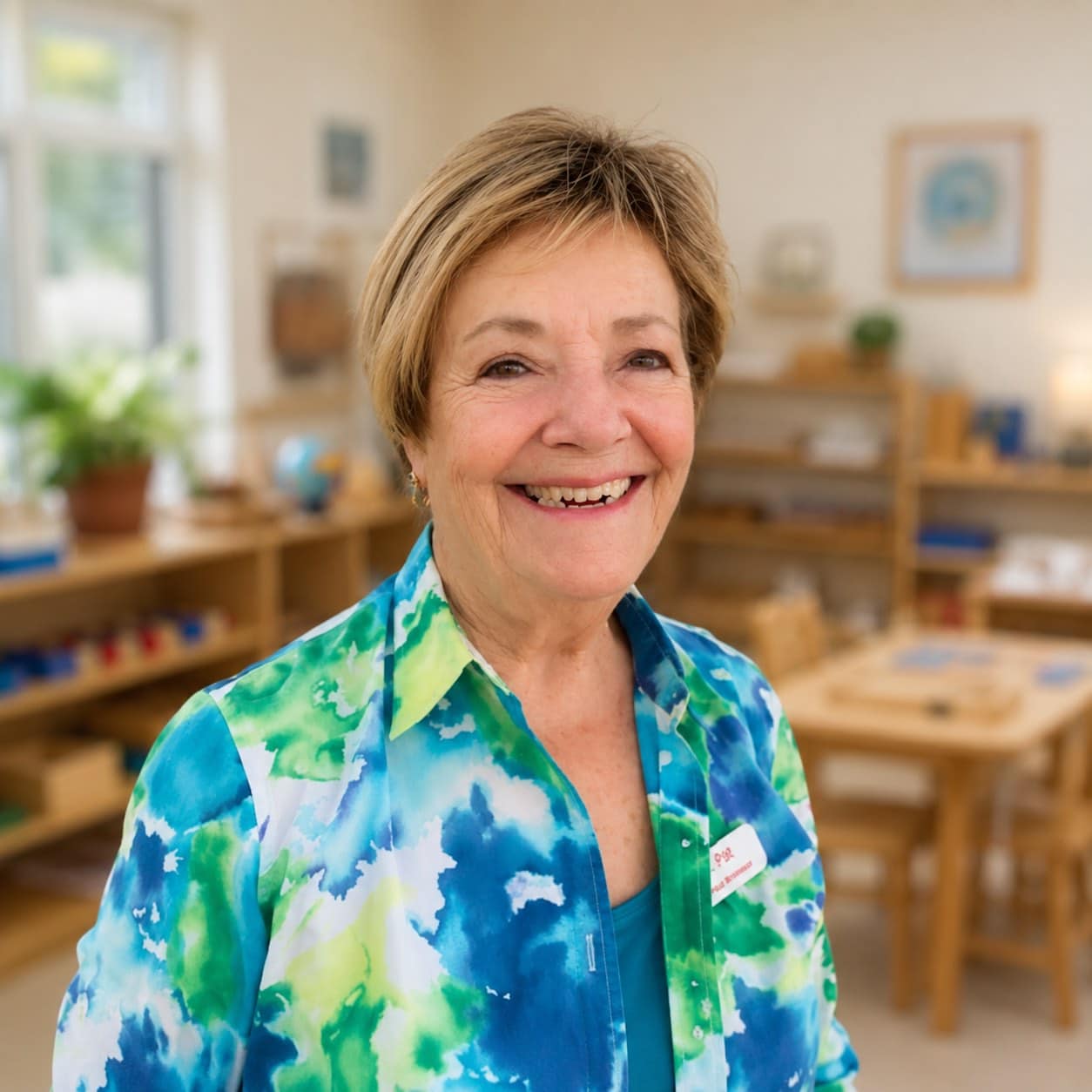 A smiling woman wearing a blue and green patterned shirt with a name tag, standing in a classroom.